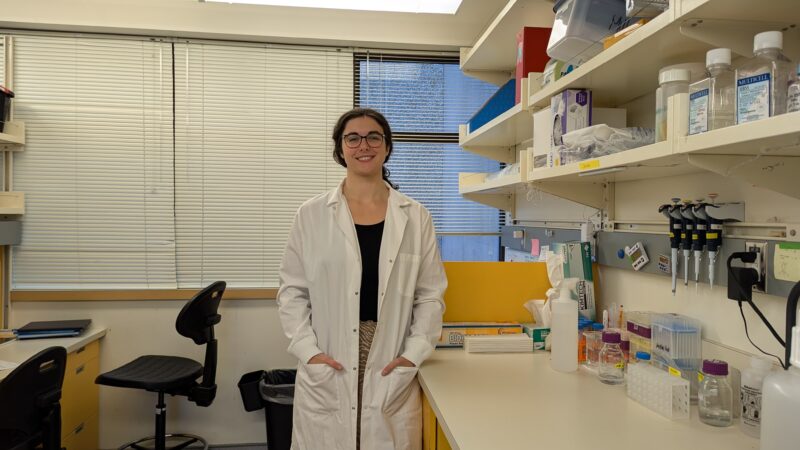 PhD candidate Sarah Ebert smiles to camera, wearing a white lab coat and standing next to her work bench in the Miller lab.